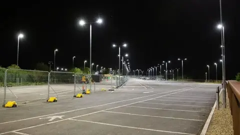 Mike Barrett A large car park with multiple street lights, shown at night with the lights on and a barrier down the middle