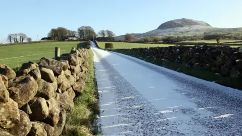 Pacemaker A country lane covered in snow runs through the middle of stone walls and fields. Slemish mountain is in the top right of the picture 
