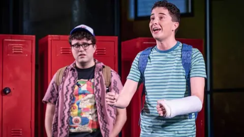 Marc Brenner Tom Dickerson (left) has brown curly hair and glasses. He is wearing a T-shirt with a large multi-coloured design on a black background and a pink shirt with a darker pink pattern on it. He has his hands in his pockets and is looking at Evan with concern. Ryan Kopel (right) has short brown hair and wears a turquoise T-shirt with a darker blue horizontal stripe. He is singing and hold his arms out, the left one has a cast on it.