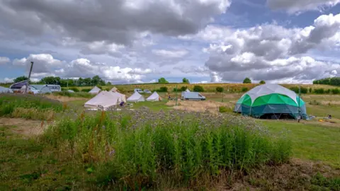 Supplied A number of tents are in the middle of the photo, including the largest one, which is white and green. A van is also in the centre and the sky, including grey clouds, features in about half of the image.