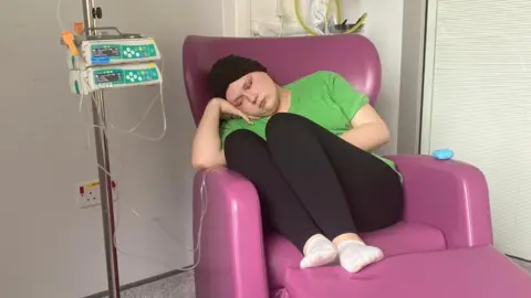 Grace and Dan Ashwell A teen girl sitting in a pink chair wearing black legging and a green t shirt. She has a black scarf around her head and is sat attached to wires in hospital. 