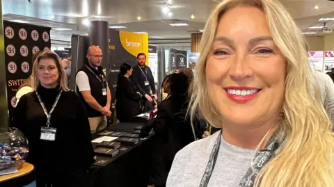 Angela Edwards smiles into the camera. She has long blonde hair and is wearing pink lip stick, and a grey jumper. Behind her business schools are set up with students visiting them.