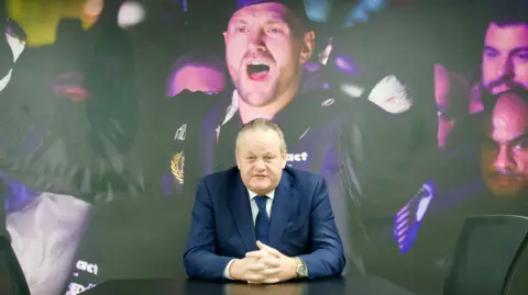 Iain McCallister sits in his boardroom, with a giant photograph of Tyson Fury covering the wall behind him. He is wearing a blue suit, white shirt and blue tie and is sat a table with his hands clasped together.