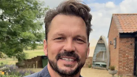 Jimmy Doherty smiles at the camera. He has short brown hair and a brown beard. He wears a blue shirt.