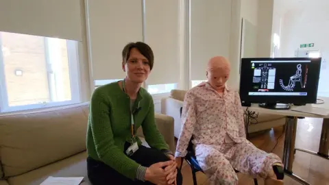 Occupational therapist Ruth Alecock sitting in a room next to robot Mona. There is a screen behind them which shows impact of touch on the robot. 