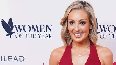 PA Media Amy Dowden in a red sleeveless trouser suit in front of a 'Women of the Year' poster. Amy has shoulder length blonde hair and is wearing oversized gold hoop earrings and a gold necklace