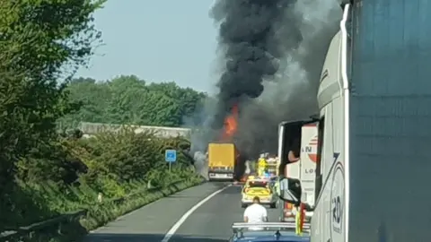A yellow lorry pulled up on to the hard shoulder of the M20 motorway has flame and dark, black smoke emerging from the vehicle. Traffic is at a standstill on the motorway. Kent Fire and Rescue Service and Kent Police are in attendance.