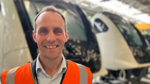 A man in an orange hi vis vest is stood in front of three trains.