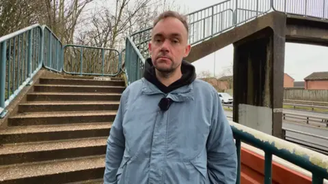 Man in blue coat stood in front of stone steps leading to bridge crossing large road