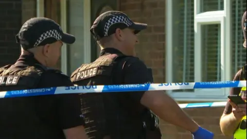 Two officers in uniform are seen working on the case outside a house, behind police tape.