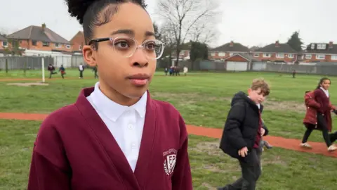 Alaia has her dark hair tied into a bun, she is wearing clear plastic glasses with black arms. She is wearing a burgundy Ferndale Primary School cardigan and a white polo shirt. She is standing on a grassy school field which has an orange-surfaced running track and fellow pupils can be seen running behind her. 