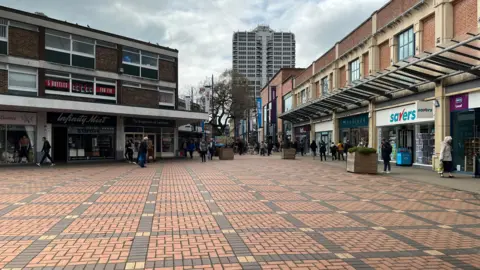 A view of Swindon town centre. There is a large paved area with two rows of shops either side. The area looks tired. In the distance is the John David Murray tower block.