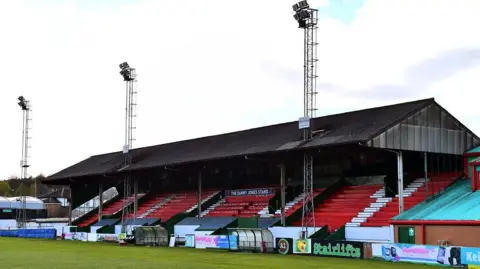LDRS A rugby club stand with red seats and a black roof. There are advertising hoardings around the bottom and three tall posts with floodlights on.