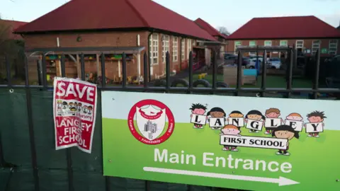 A general view of the entrance to Langley First School. On the railings outside there is a large banner pointing to the school's entrance and a poster saying Save Langley First School.