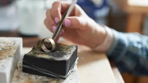 A jeweller maker using tweezers to work on a ring. The hand and arm of the female jeweller can be seen, revealing pink painted nails and a blue shirt.