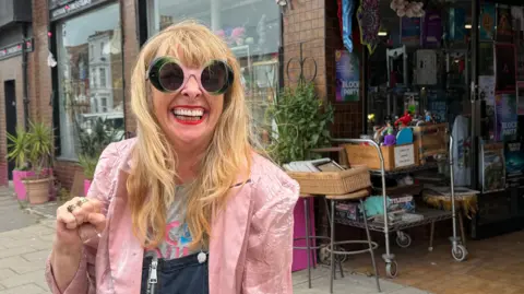 A woman with long blonde hair, pink and green glasses and pink jacket.