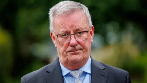 PA Media Mike Nesbitt has grey hair. he is wearing black rimmed glasses and looks sternly at the camera. He is wearing a dark suit, light blue shirt and silver and white patterned tie.