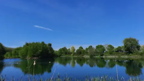 BBC Weather Watchers/Ruckynloo A view of a lake with trees in the distance reflected in the water including the pure blue sky overhead. On the water, a couple of ducks can be seen in the distance gliding over it.