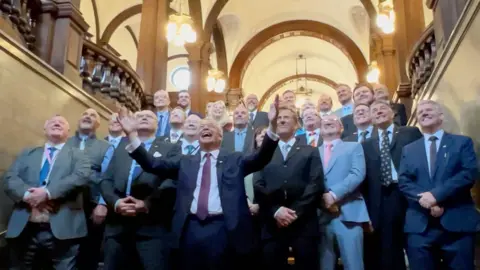 Nige Farage pictured with his arms apart and raised and smiling. There are more than 20 councillors behind him. Most are wearing suits and they are smiling and several men at the front have their hands folded in front of them.
