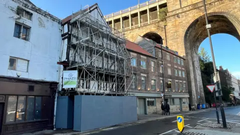 The Cooperage completely covered in scaffolding. A sign of a scaffolding firm is attached on the side of the building. Two people are walking past on the pavement. A row of three and four-storey brick houses is next to The Cooperage. The High Level Bridge is rising above the buildings. 