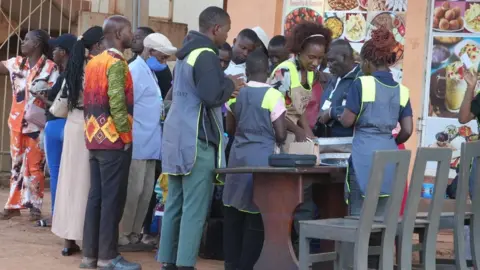 A group of voters stands in line outdoors as election officials in reflective vests assist with voter verification at a table outside a building decorated with food posters 