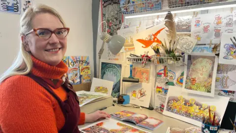 An artist sits at her desk surrounded by watercolour paintings that feature in her Adventure Mice books. A book lays open on her desk along with pots of pencils.