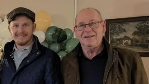 Family photo A man with a light brown beard, a flat cap and a blue coat standing in a room with balloons behind him, alongside an older man with thinning grey hair, black framed glasses and a green coat