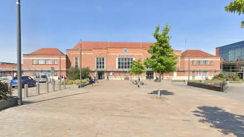 Google Exterior of Doncaster Railway Station, a redbrick building with a clock at the centre of its facade, which looks out over a pedestrian area, dotted with trees.