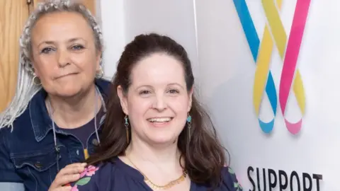 Steffi Colbert and Nat are both looking at the camera and smiling. Steffi stands to the left of the image, just behind Nat. She has grey dreadlocks in a half up half down hairstyle and wears a dark blue denim shirt with a deep purple cotton round neck t shirt underneath, her hand is resting on Nat's shoulder. Nat has dark brown/red hair which is also in a half up do. She wears turquoise pendant earrings and a gold necklace, with a navy blue blouse which has a pink floral print on the shoulder. To the right of the image is a large sign for Wessex Cancer Support, which is a 'W' in the shape of two looped pieces of ribbon in a mixture of blue, yellow and pink colours.