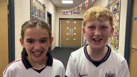 Scarlett, a girl with brown hair, and Henry, a boy with ginger-blonde hair and freckles, cheering on England in their white football shirts. They are standing in a school corridor which has St George flag bunting as well as the noticeboards and images common in primary schools. Double doors behind them lead to the nursery and lower school.