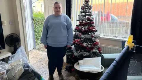 Hannah McCann stands in her flooded home beside her Christmas tree.  The floor is covered in mud.  She is wearing a blue jumper, dark leggings and brown boots.   A baby's cot is in the foreground. 