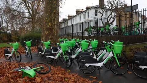 Reuters A large number of Lime bikes parked on a path in a park with others behind a fence next to a road. There are trees in the park and rows of houses in the background