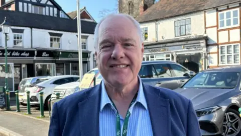 There is a man in a dark blue suit jacket, a light blue shirt with white pinstripes and his top button undone. He is wearing a dark green lanyard with 'Church Stretton Town Council' written on logos around the flag. He also has grey hair. He is stood in front of a car park. 