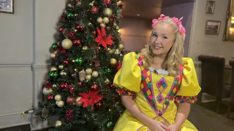 Lucy Conley in a yellow goldilocks dress with diamond detail in red, green and yellow. There is a Christmas tree with red ribbons and gold baubles behind her. Ms Conley is also wearing pink ribbons in her hair.