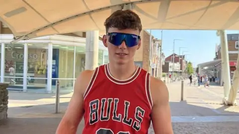 A young man with curly brown hair and sunglasses wearing a red vest with the word "BULLS" on it. He is stood in front of a high street on a sunny day.