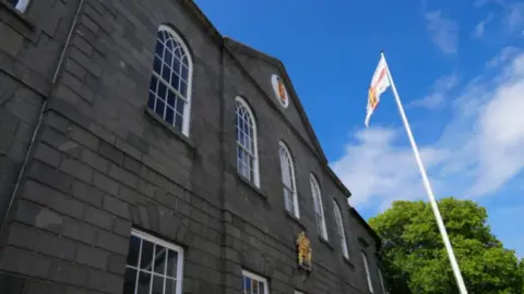 A wide shot of the States of Guernsey building. It is a dark grey stone building with the Guernsey flag raised on a pole in front of the building.