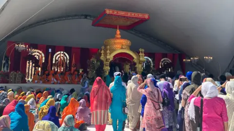 A large crowd sits and stands beneath a white canopy facing a raised stage. On the stage, an ornate golden structure with red drapery and chandeliers forms the backdrop, with several people in orange robes seated in front of it. The audience is dressed in colourful traditional clothing and head coverings, filling the space from the foreground to the stage.