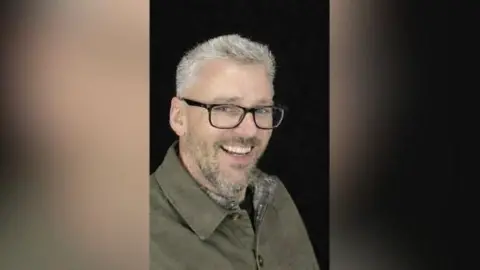 Avon and Somerset Police Craig Hurcombe smiles at the camera. He wears a green collared jacket and black rimmed glasses. He has short grey hair and a beard. 