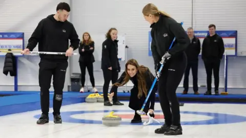 Catherine crouches down to push a curling stone across the rink, with two members of staff standing at either side.