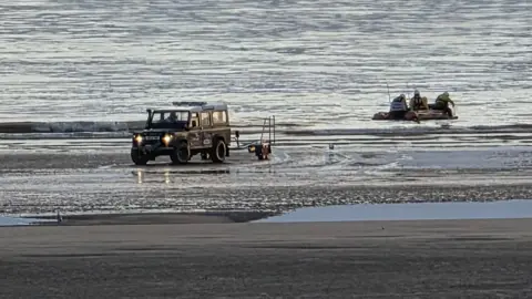 A 4x4 vehicle waits at the shore for an inflatable RNLI lifeboat with crew members in it in the sea.