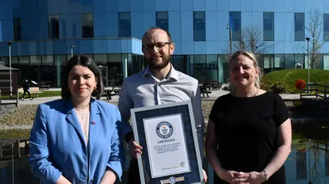 Royal Papworth Charity Alex with dark hair and beard, wearing a light=coloured shirt and holding a certificate in a frame, is flanked by Ms Grant, who has medium-length dark hair and is wearing a blue jacket, and Shelley Green, with long blonde hair wearing a black T-shirt. They are standing in front of a hospital building which has mainly glass on the ground floor and a blue wall with narrow windows on the first floor.