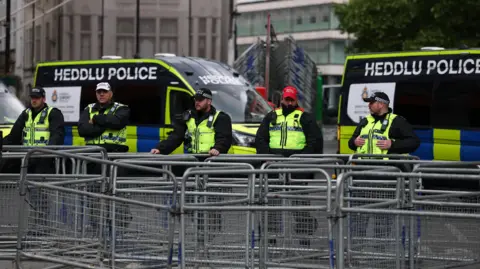 Reuters Police officers stand behind a row of metal barriers in front of two police vans which say HEDDLU POLICE in dual Welsh and English signage.