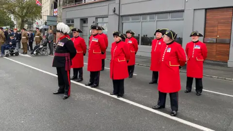 A group of eight Chelsea Pensioners wearing their red tunics and military medals stand on a road in front of a grey building in Guernsey. Another man wearing a black military suit and hat with white feathers on top of it and military medals on his chest is stood in front of them.