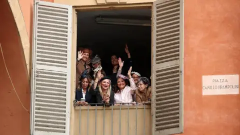 Reuters A group of women wearing fascinators and hats wave out of a shuttered balcony on a terracotta-walled building