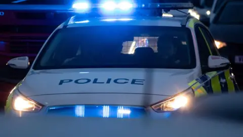 Two police officers drive a police car with its blue lights flashing through traffic in the dark in Bristol