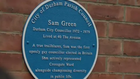 A round blue plaque with white writing saying Sam Green was a Durham County Councillor from 1972 to 1979 and lived at 40 The Avenue. It also says that Green was a true trailblazer and the first openly gay councillor to be elected in Britain and that he represented the Crossgate Ward. 