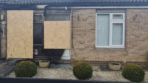 A brick-built bungalow with three small ornamental shrubs in a gravel garden in front has two large pieces of chipboard covering blackened, burnt-out windows.