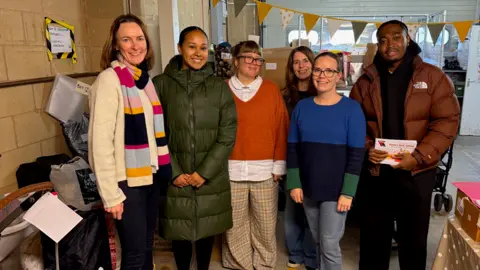 Baby Bank Network Five people from the network are joined by Gamba Cole standing in a warehouse by boxes of donations. There is yellow and white bunting in the background
