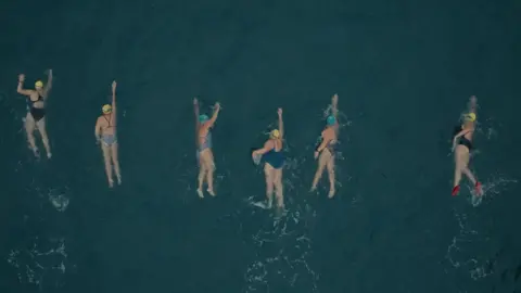Braving the Blue - Our Lives, BBC Cymru Wales Six women swimming in the dark blue sea. All are wearing swimming costumes, swim caps and goggles. One of the women also wears red swim shoes. 