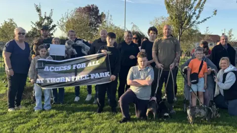 About two dozen people are standing in a park and facing the camera, with some pulling defiant poses. They are holding up signs that say "Access for all at Thorntree Park".